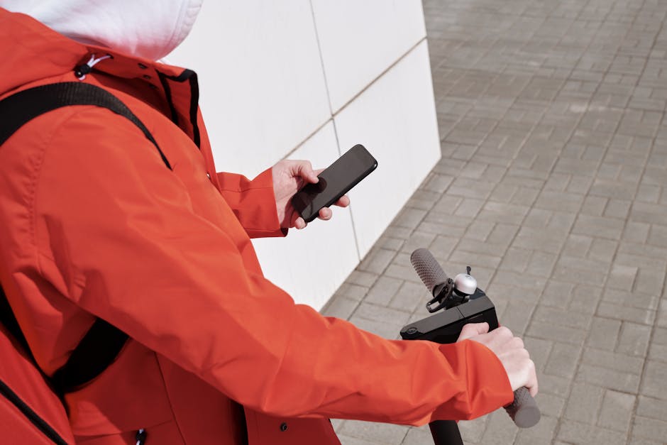 Person in red jacket using smartphone while riding a scooter outdoors.
