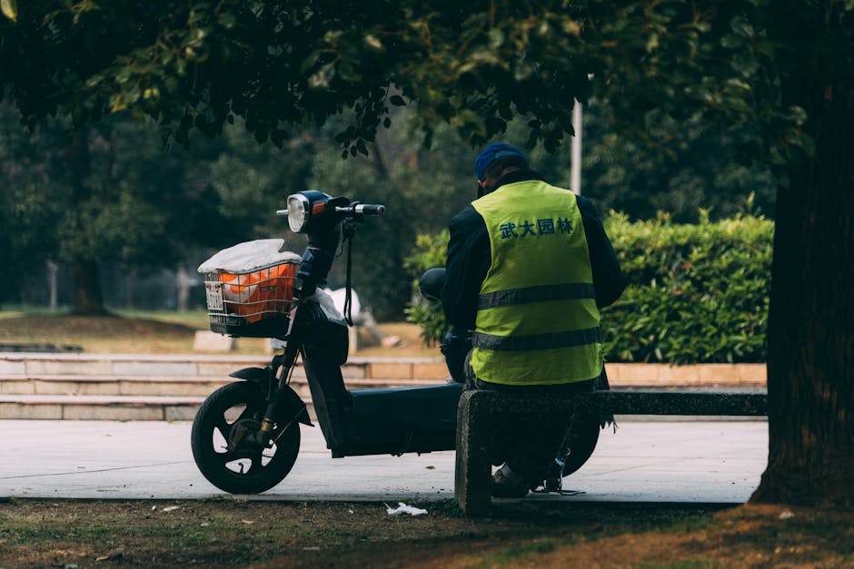 A man in a reflective vest sits on a bench next to a scooter in a city park.