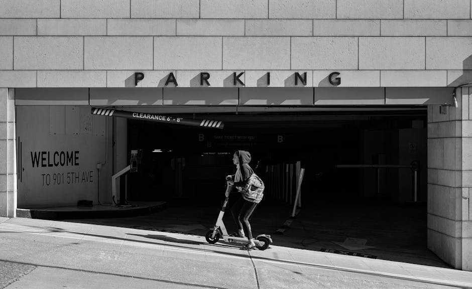 Young woman riding an electric scooter past a parking garage entrance in grayscale.
