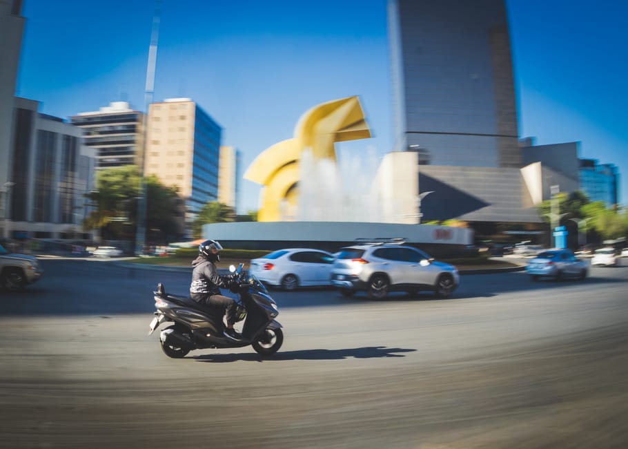 A scooter rider swiftly passing by a modern cityscape with a prominent yellow sculpture.