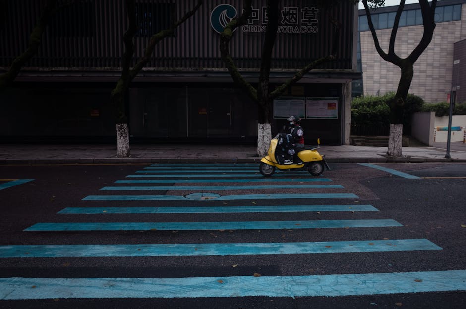 A lone rider on a yellow scooter moves through an urban crosswalk at dusk.